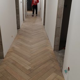 Gut-pro usługi remontowe - Korytarz w trakcie wykończenia z ułożoną podłogą z płytek imitujących drewno w jodełkę. Widoczne surowe ściany i otwory drzwiowe. Osoba w oddali.