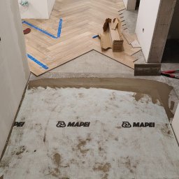Gut-pro usługi remontowe - Układanie parkietu w jodełkę w korytarzu, widoczna warstwa hydroizolacji Mapei, narzędzia glazurnicze i częściowo ułożona podłoga. Prace wykończeniowe w trakcie.