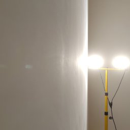 Gut-pro usługi remontowe - Świeżo pomalowana ściana w rogu pokoju oświetlona przez żółty reflektor budowlany. Widoczne niedoskonałości powierzchni pod światło.