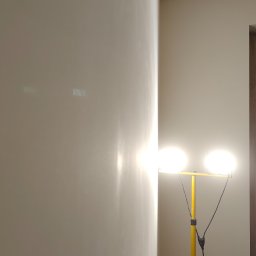 Gut-pro usługi remontowe - Gładka, świeżo pomalowana ściana w jasnym odcieniu, oświetlona lampą budowlaną na żółtym statywie. Widoczny fragment drzwi i sufitu. Minimalistyczne wnętrze po malowaniu.