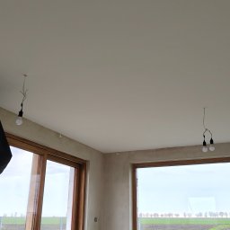 Gut-pro usługi remontowe - Surowy stan wnętrza z dużymi oknami i widokiem na zewnątrz. Ściany przygotowane do malowania, widoczne kable oświetleniowe. Podłoga betonowa, brak wykończenia.