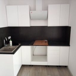 Gut-pro usługi remontowe - Nowoczesna, minimalistyczna kuchnia z białymi szafkami i czarnym blatem. Widoczny zlew, bateria i okap. Część szafek jeszcze nie posiada frontów.