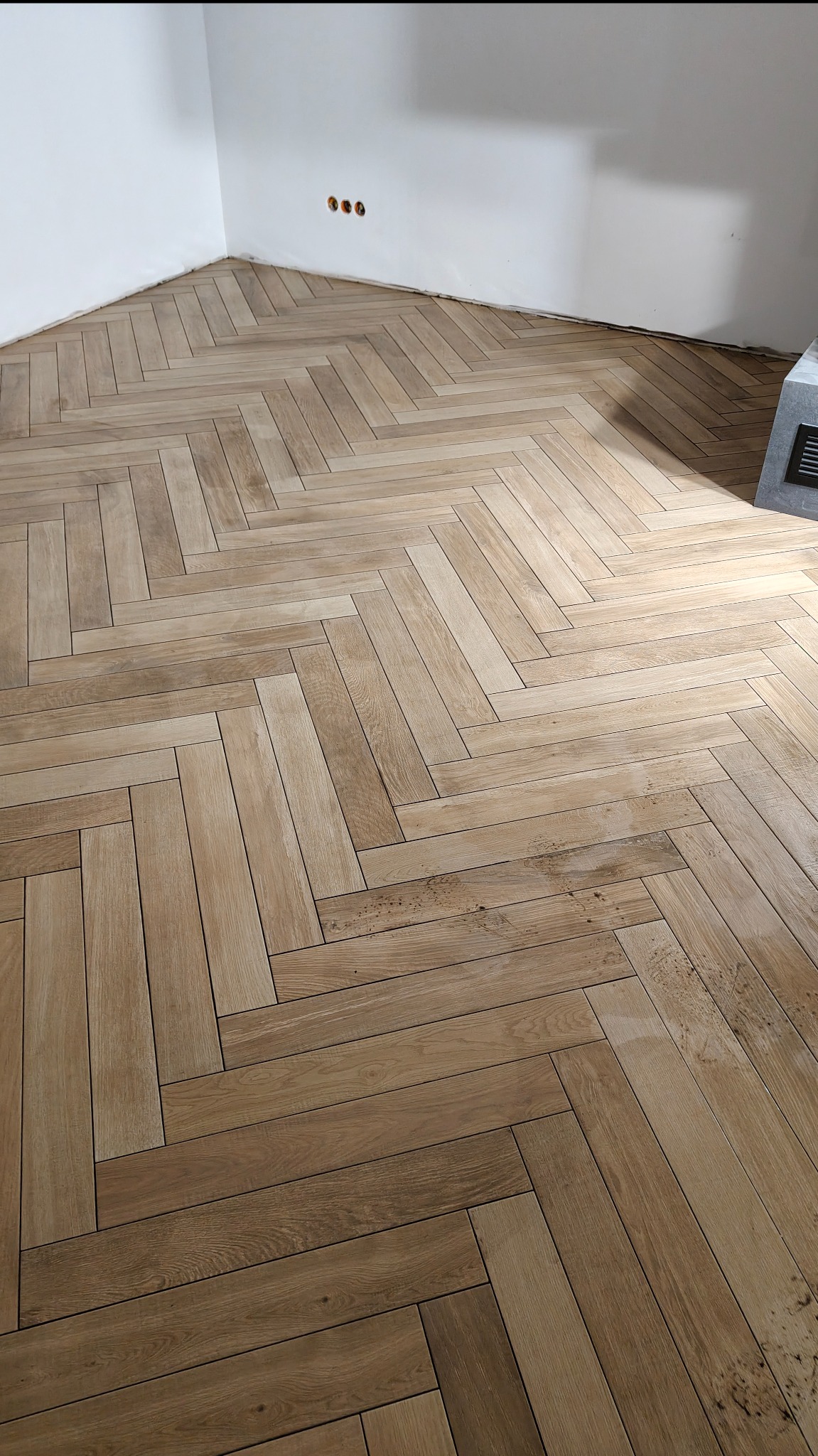 Ukończona podłoga z płytek imitujących drewno, ułożonych w jodełkę, w rogu pomieszczenia z widocznymi gniazdkami elektrycznymi i fragmentem grzejnika.