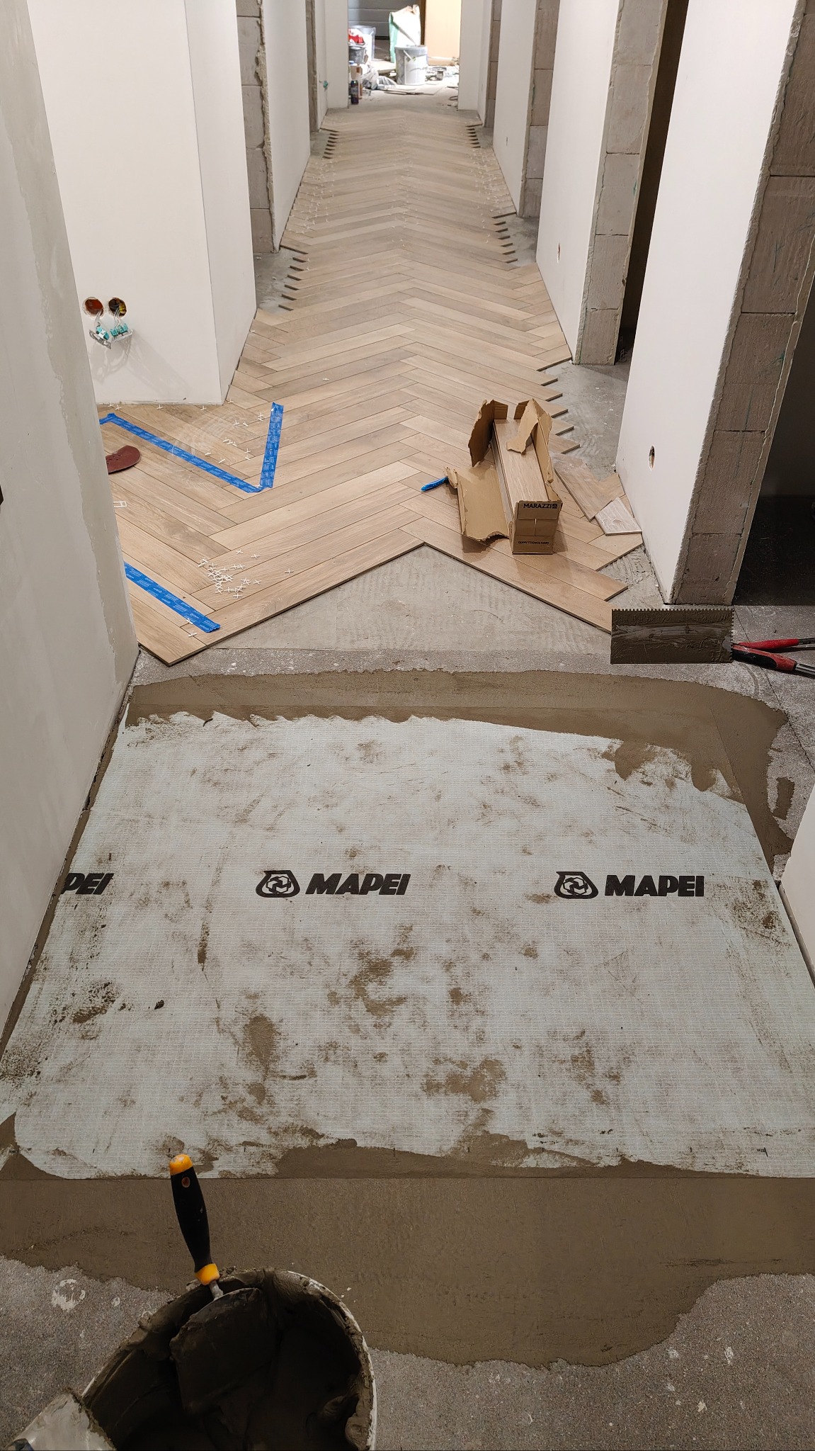 Układanie parkietu w jodełkę w korytarzu, widoczna warstwa hydroizolacji Mapei, narzędzia glazurnicze i częściowo ułożona podłoga. Prace wykończeniowe w trakcie.