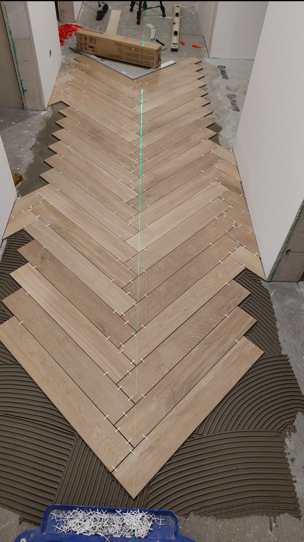 Układanie parkietu w jodełkę z użyciem lasera i krzyżyków dystansowych. Widoczna warstwa kleju i narzędzia glazurnicze. Jasne drewno, precyzyjne wykonanie.