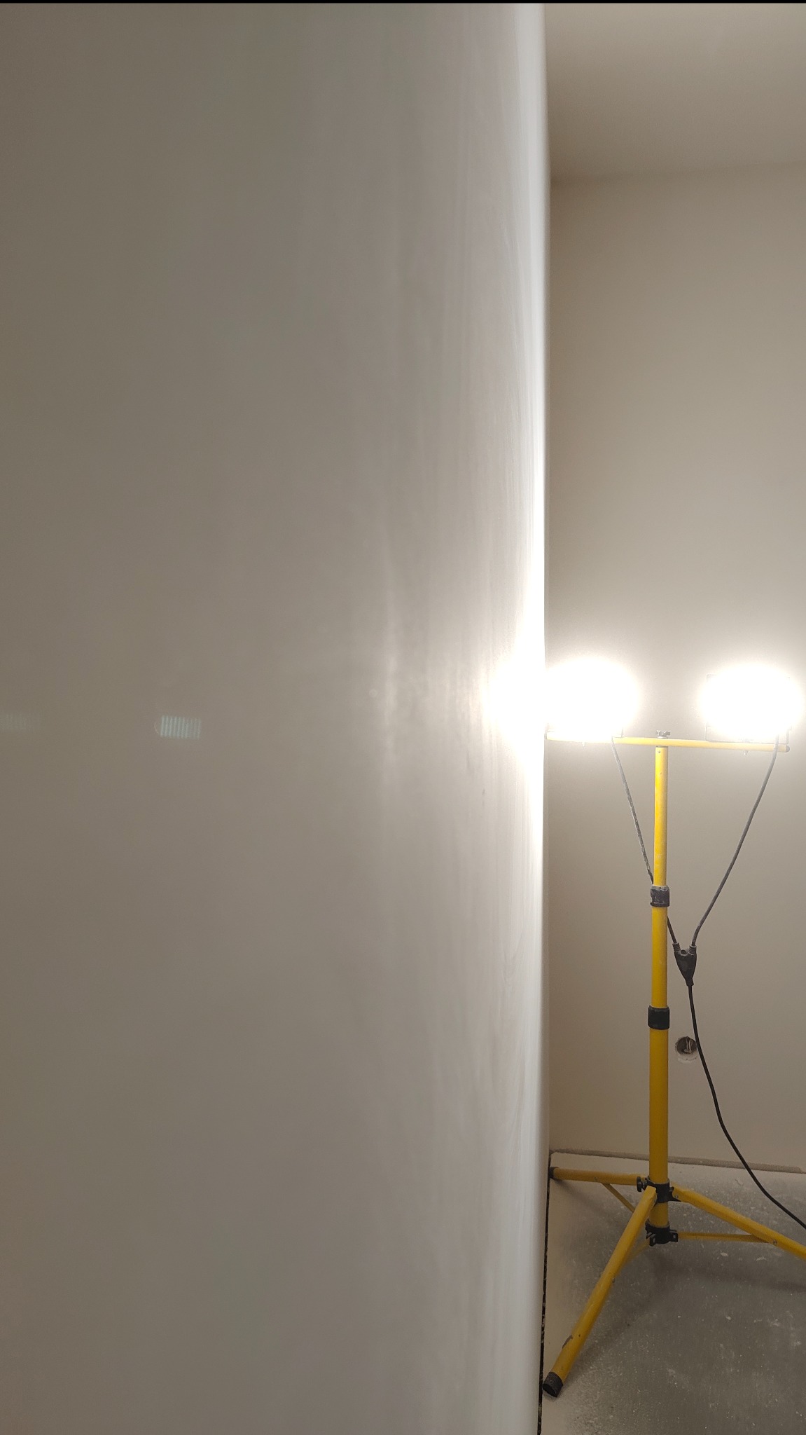 Świeżo pomalowana ściana w rogu pokoju oświetlona przez żółty reflektor budowlany. Widoczne niedoskonałości powierzchni pod światło.