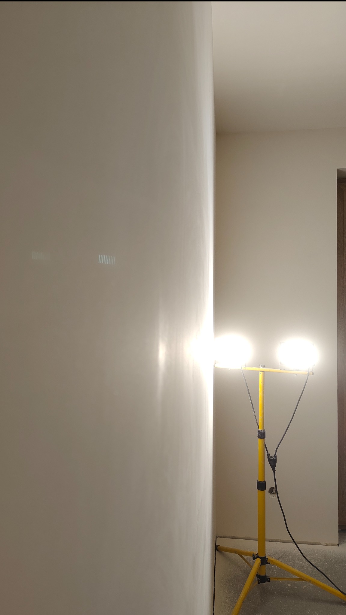 Gładka, świeżo pomalowana ściana w jasnym odcieniu, oświetlona lampą budowlaną na żółtym statywie. Widoczny fragment drzwi i sufitu. Minimalistyczne wnętrze po malowaniu.