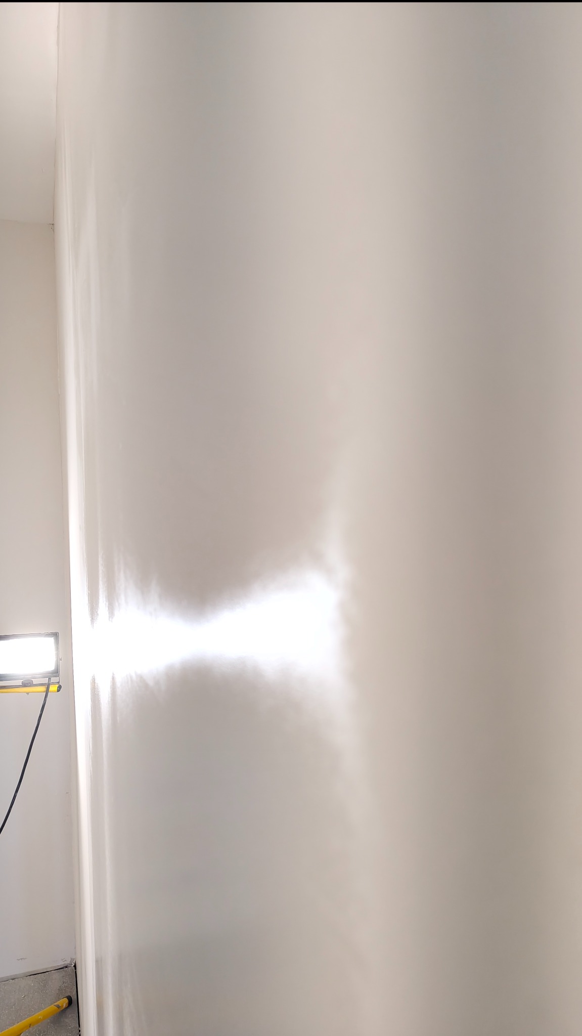 Świeżo pomalowana, gładka ściana w jasnym odcieniu bieli, oświetlona lampą budowlaną, ukazująca precyzję wykonania i brak niedoskonałości. Widoczne ślady po wałku.