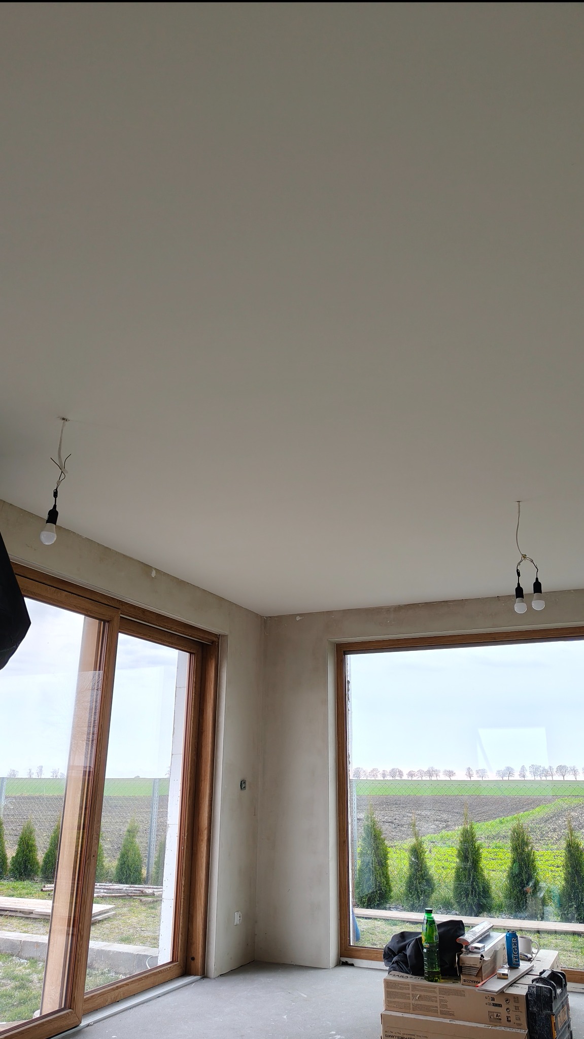 Surowy stan wnętrza z dużymi oknami i widokiem na zewnątrz. Ściany przygotowane do malowania, widoczne kable oświetleniowe. Podłoga betonowa, brak wykończenia.