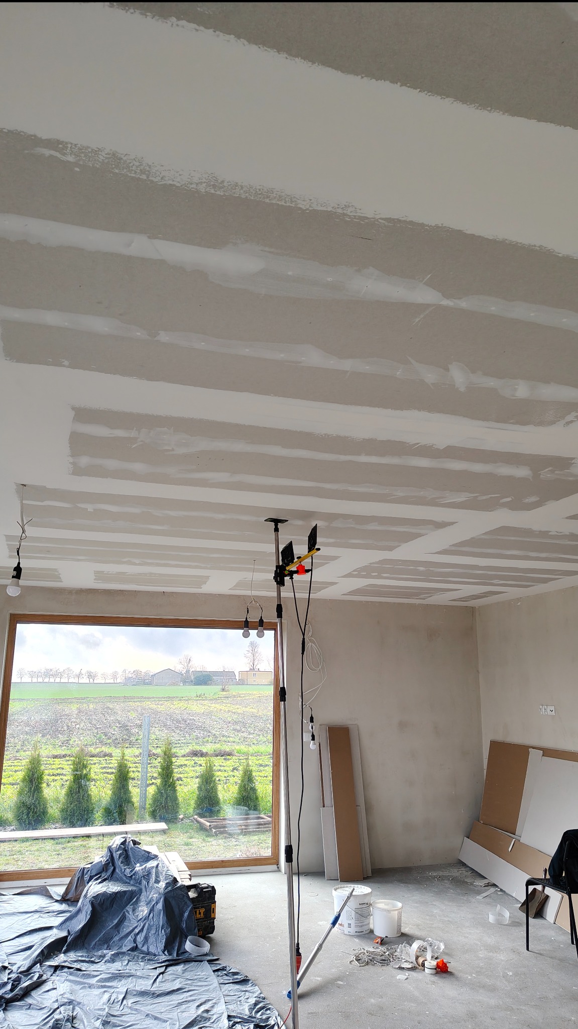 Sufit z płyt kartonowo-gipsowych w trakcie szpachlowania, widoczne łączenia. W tle okno z widokiem na zieleń i narzędzia pracy. Oświetlenie budowlane.