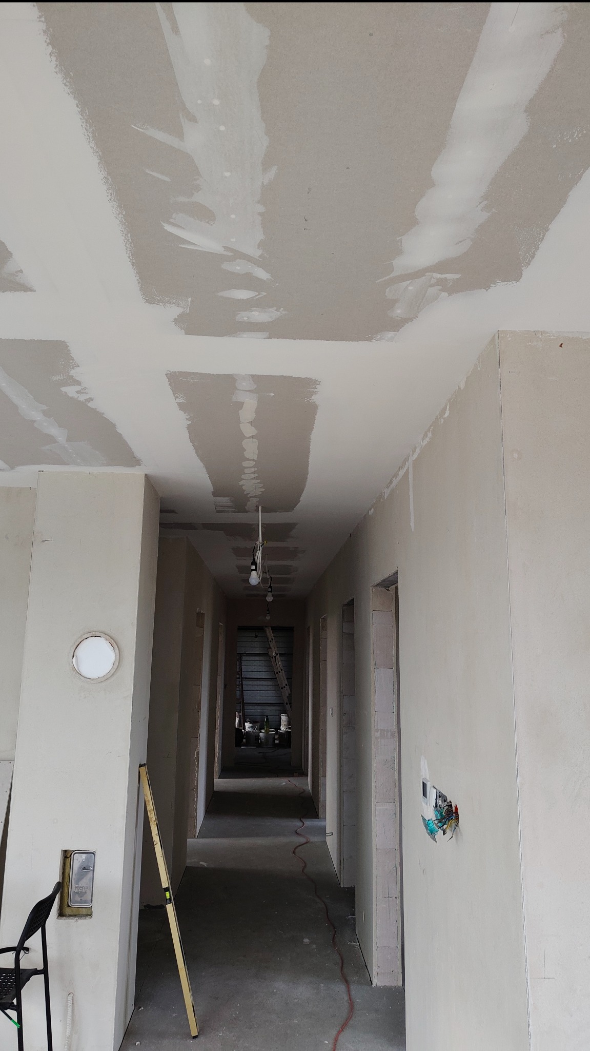 Korytarz w trakcie szpachlowania płyt karton-gips na suficie i ścianach. Widoczna drabina w głębi, poziomica oparta o ścianę i wystające kable elektryczne.