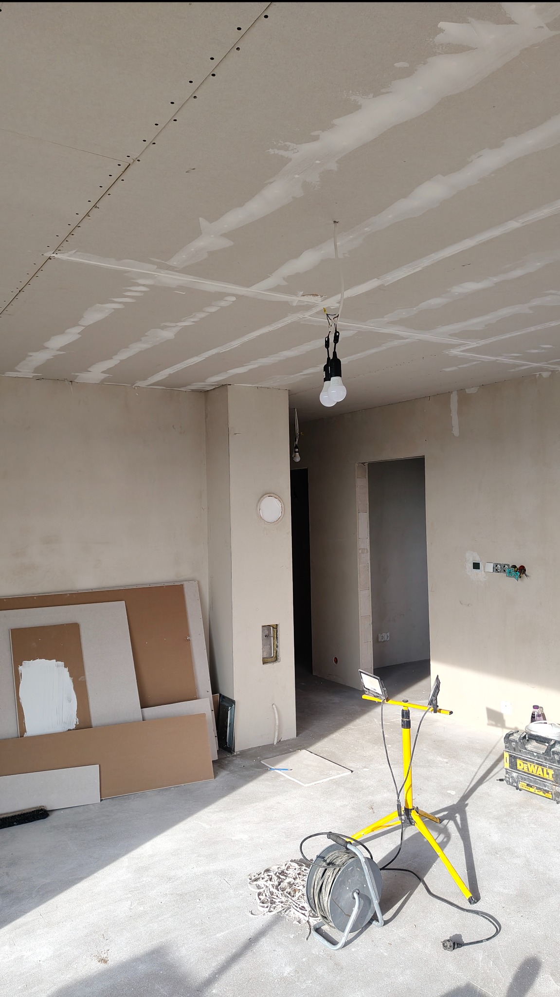 Wnętrze w trakcie prac wykończeniowych: zamontowane płyty karton-gips na suficie, widoczne łączenia szpachlowane, lampa budowlana, płyty gipsowe i narzędzia na podłodze.
