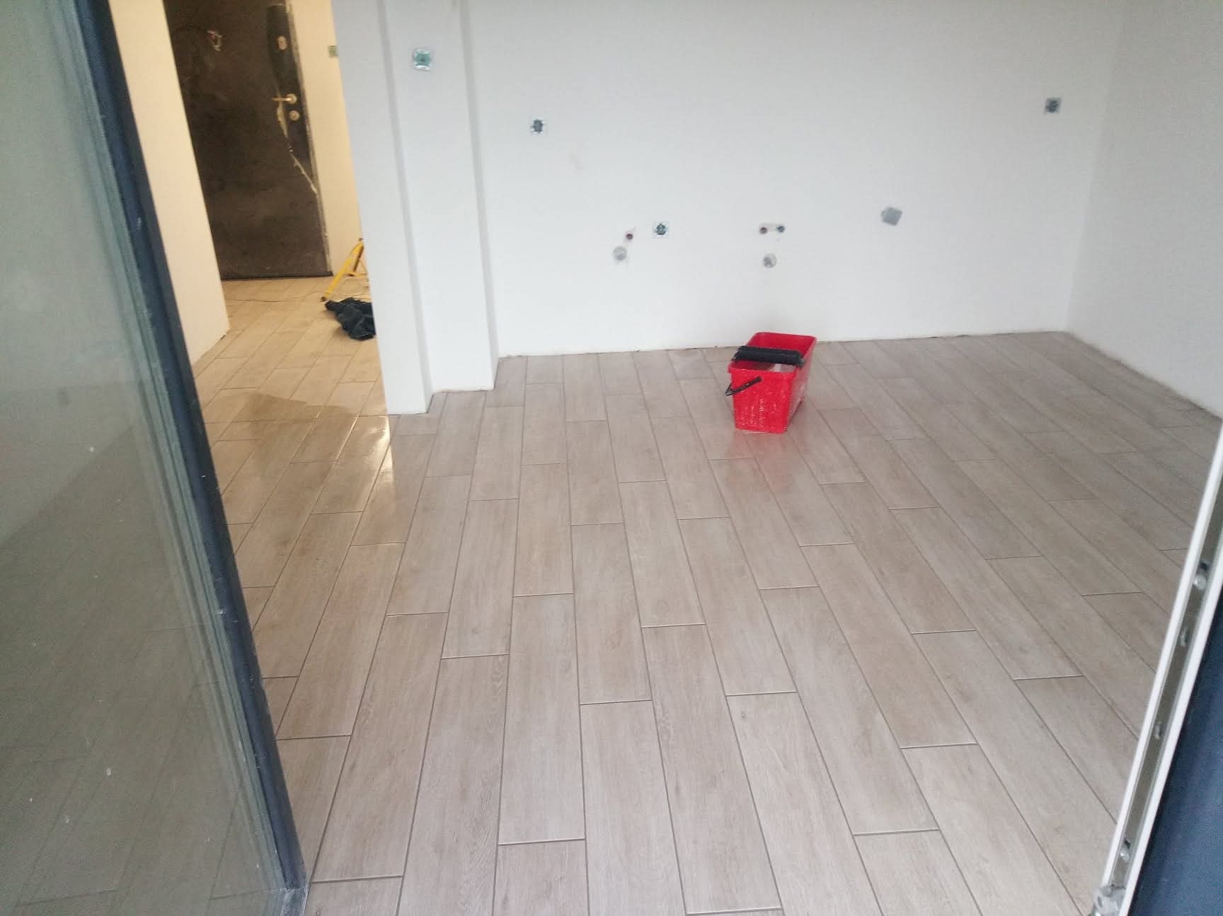 Ułożona podłoga z płytek imitujących drewno w jasnym odcieniu, widoczny czerwony pojemnik na narzędzia, białe ściany z przygotowanymi punktami instalacji.