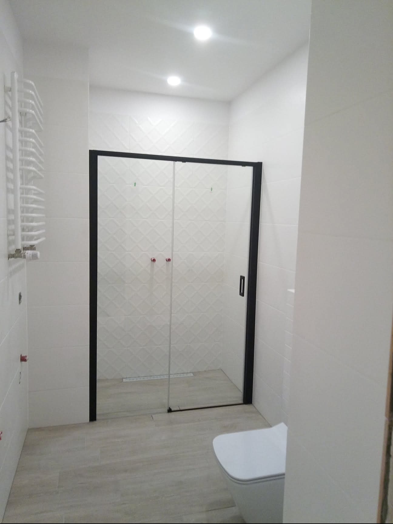 Nowoczesna kabina prysznicowa z czarną ramą i szklanymi drzwiami w jasnej łazience z białymi płytkami i grzejnikiem drabinkowym.