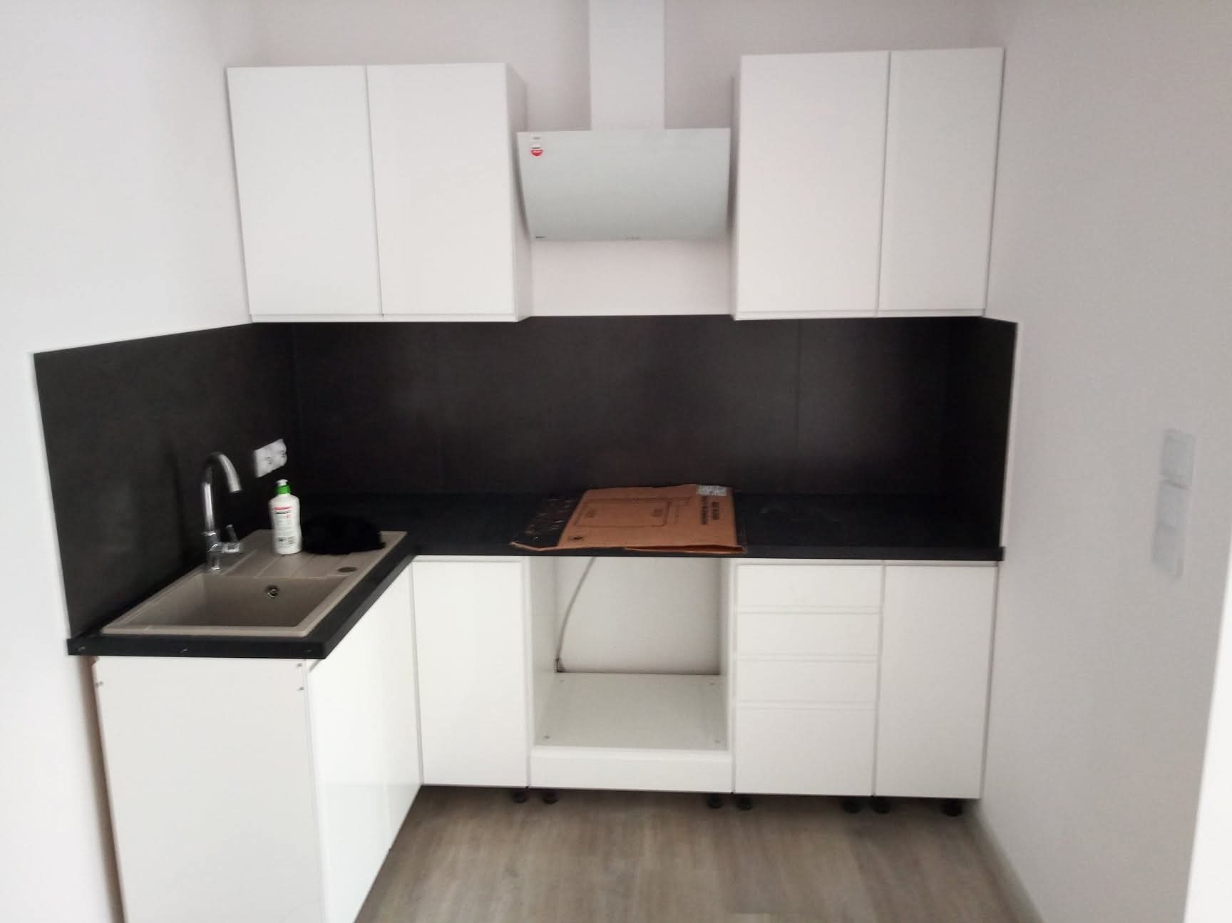Nowoczesna, minimalistyczna kuchnia z białymi szafkami i czarnym blatem. Widoczny zlew, bateria i okap. Część szafek jeszcze nie posiada frontów.