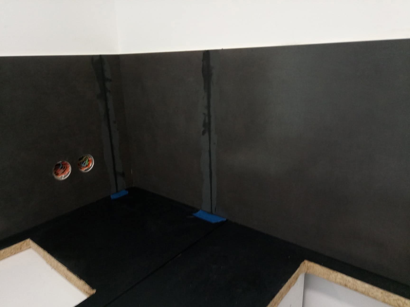 Ciemne płyty ścienne w trakcie montażu, widoczne kable elektryczne i fragment podłogi z płytami. Prace wykończeniowe w pomieszczeniu o surowym wyglądzie.