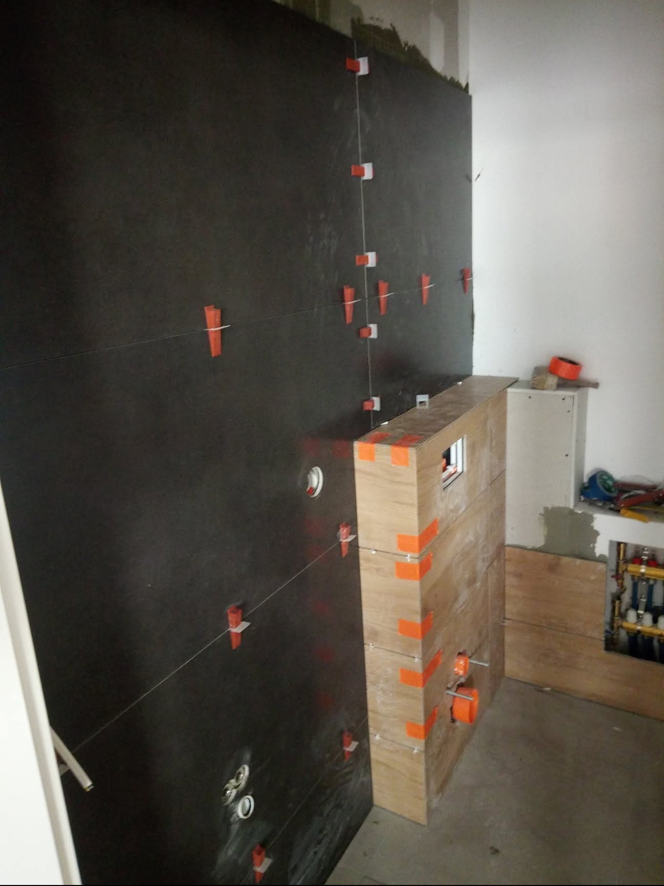 Ciemne płytki na ścianie łazienki, mocowane za pomocą klipsów poziomujących. Widoczna zabudowa z płyt drewnopodobnych i instalacja wodna w tle.