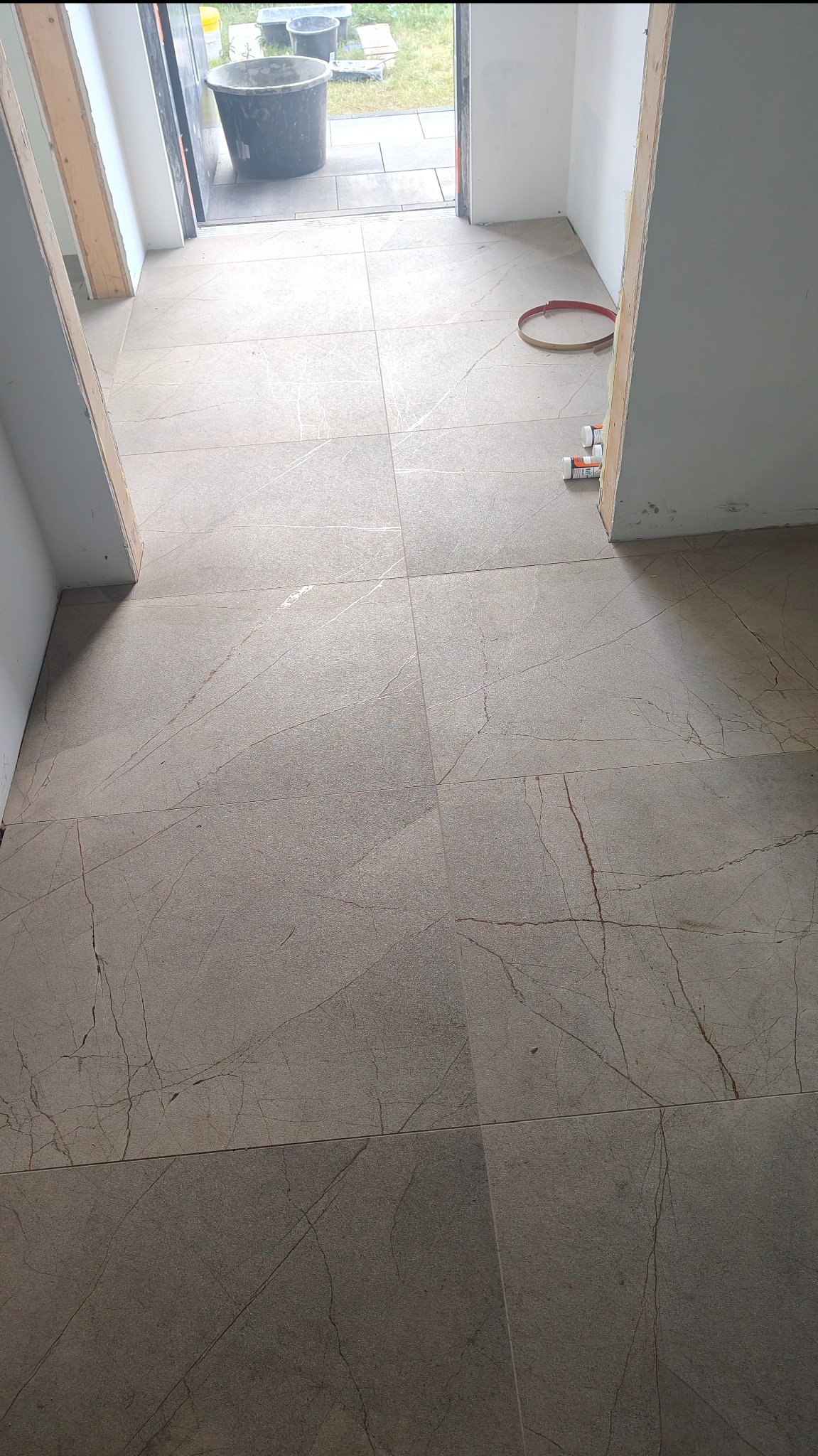 Ułożona podłoga z płytek imitujących marmur w przejściu, widoczne narzędzia glazurnicze i wiadro na zewnątrz, jasne ściany, surowy stan wykończenia.