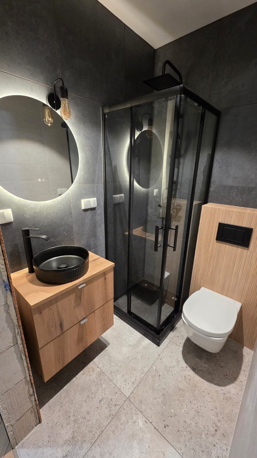 Nowoczesna łazienka z czarną armaturą: kabina prysznicowa, umywalka nablatowa na drewnianej szafce, podświetlane lustro i toaleta. Szare płytki na ścianach i podłodze.
