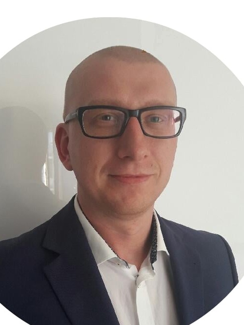 Portret mężczyzny w okularach i marynarce na białym tle. Uśmiechnięty, z krótkimi włosami. Kadr od ramion w górę, z lekkim uśmiechem.