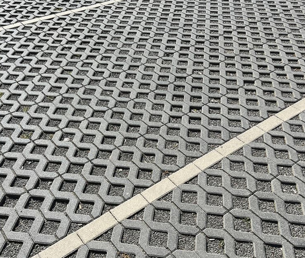 Szara, ażurowa kostka brukowa z wypełnieniem żwirowym, ułożona w regularny wzór. Widoczna linia oddzielająca fragment nawierzchni.