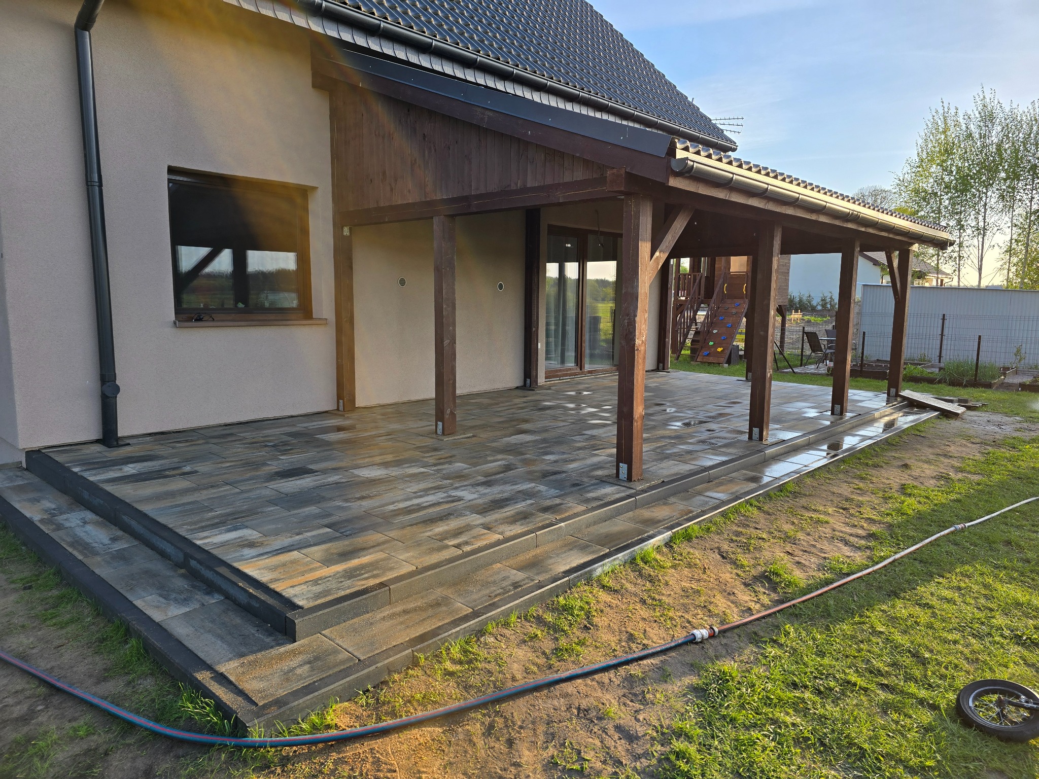 Nowoczesny taras z kostki brukowej w odcieniach szarości, przylegający do domu z drewnianą konstrukcją zadaszenia. Widoczny fragment trawnika i węża ogrodowego.
