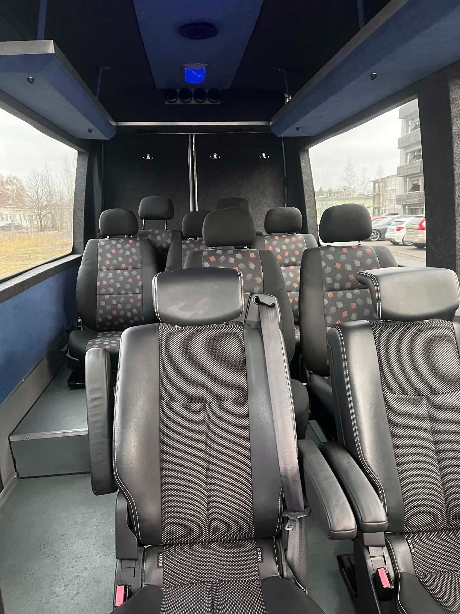 Wnętrze busa z rzędami foteli z tapicerką w geometryczne wzory. Fotele skórzane z podłokietnikami i pasami bezpieczeństwa. Widok na okno i zabudowania na zewnątrz.