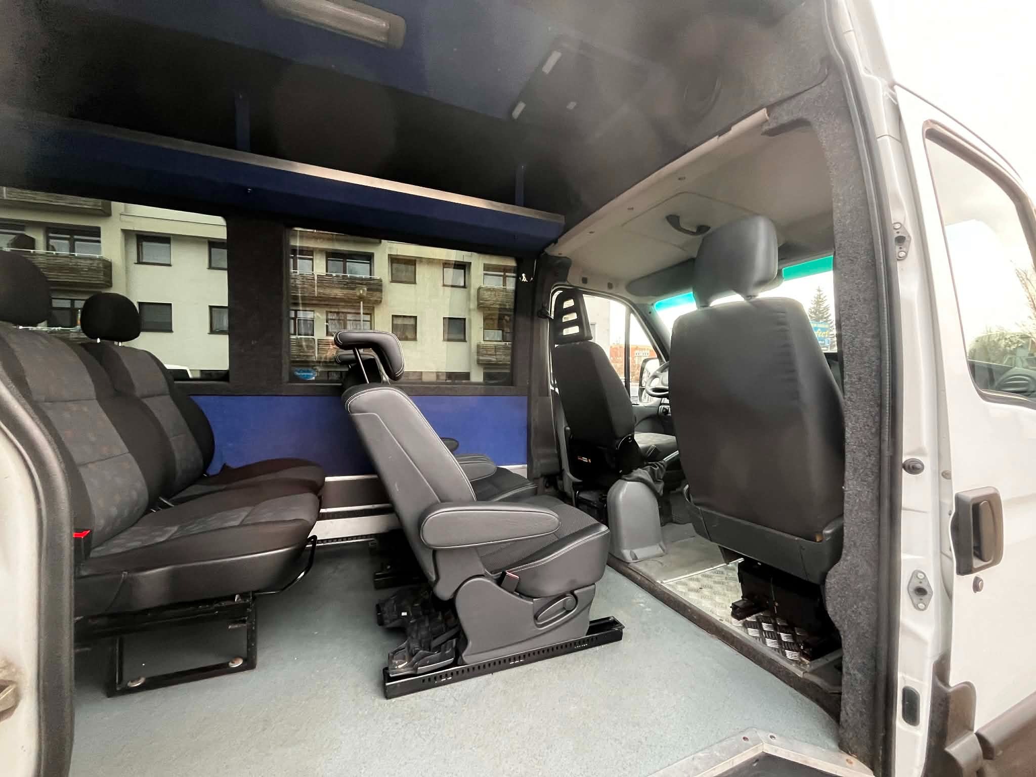 Wnętrze busa z regulowanymi fotelami pasażerskimi, widok z boku na fotele z tapicerką w odcieniach szarości i czerni, okno z widokiem na budynek mieszkalny.