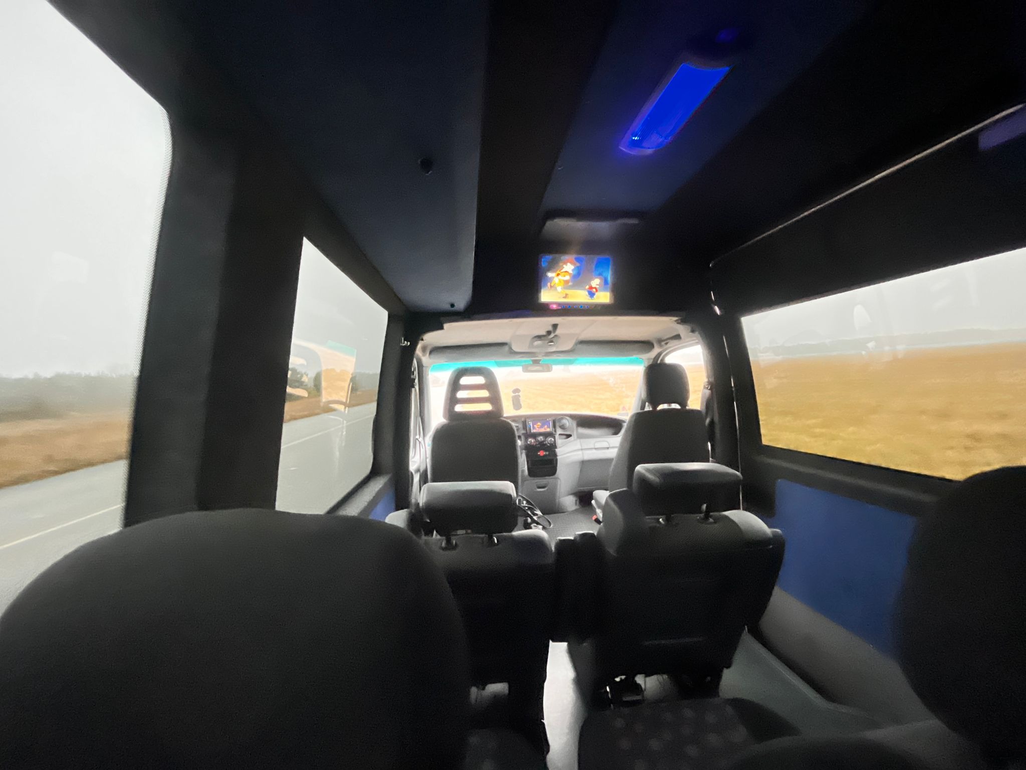 Wnętrze luksusowego busa z telewizorem dla pasażerów, widok z tyłu pojazdu podczas jazdy. Komfortowe fotele i oświetlenie LED tworzą przyjemną atmosferę podróży.