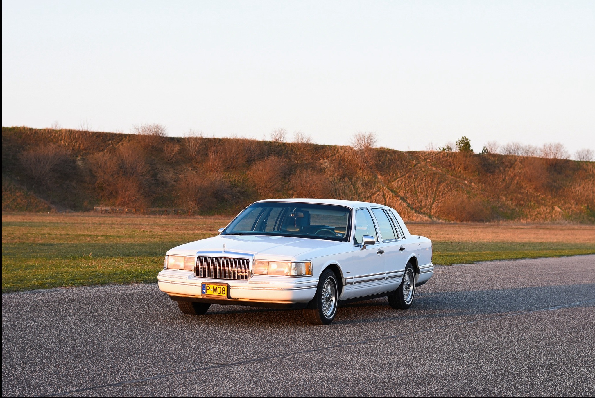 Biały Lincoln Town Car z polską tablicą rejestracyjną P W08 na tle pagórkowatego krajobrazu w ciepłym świetle zachodzącego słońca.