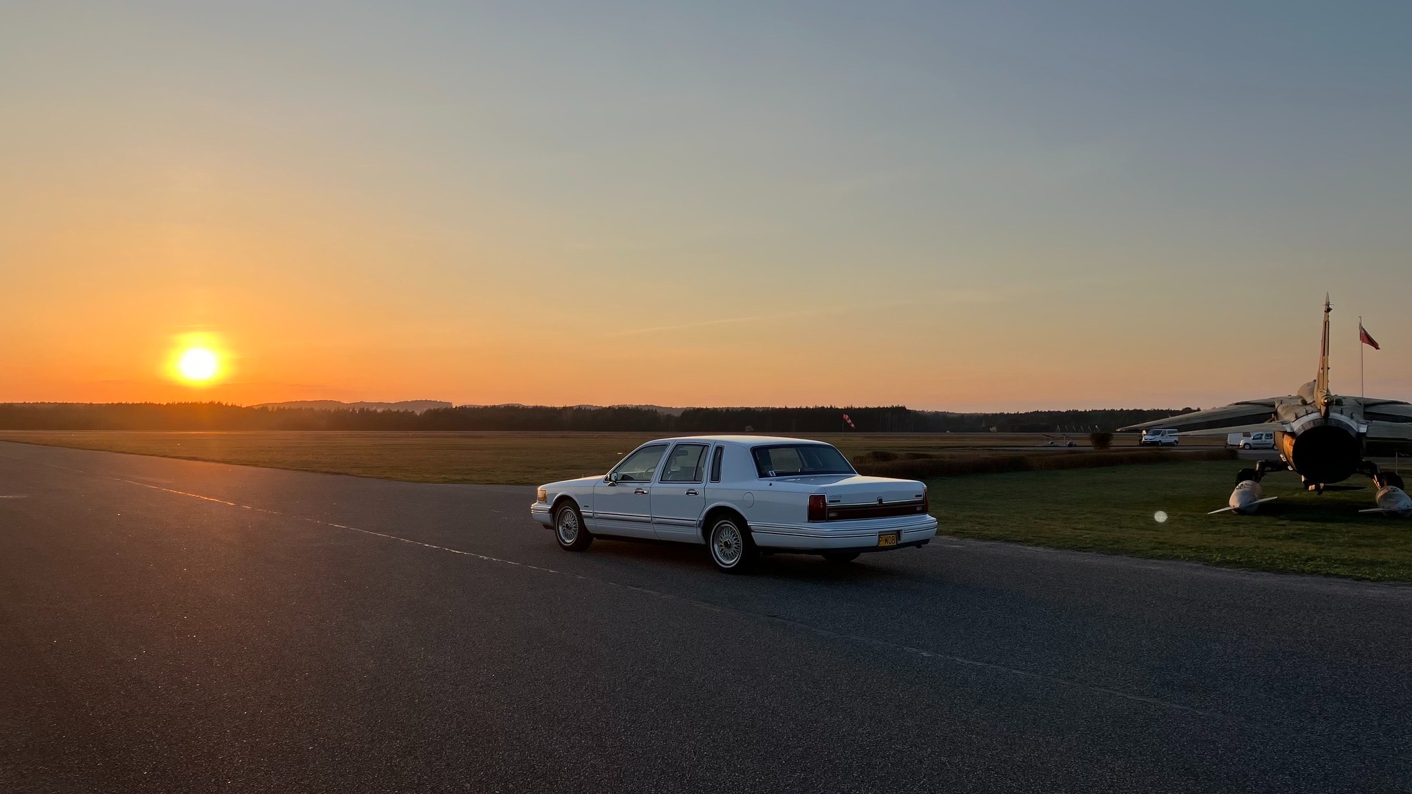 Biała limuzyna Lincoln na tle zachodzącego słońca i samolotu na lotnisku. Nietypowe połączenie luksusu i lotnictwa. Widok z tyłu, spokojna atmosfera.