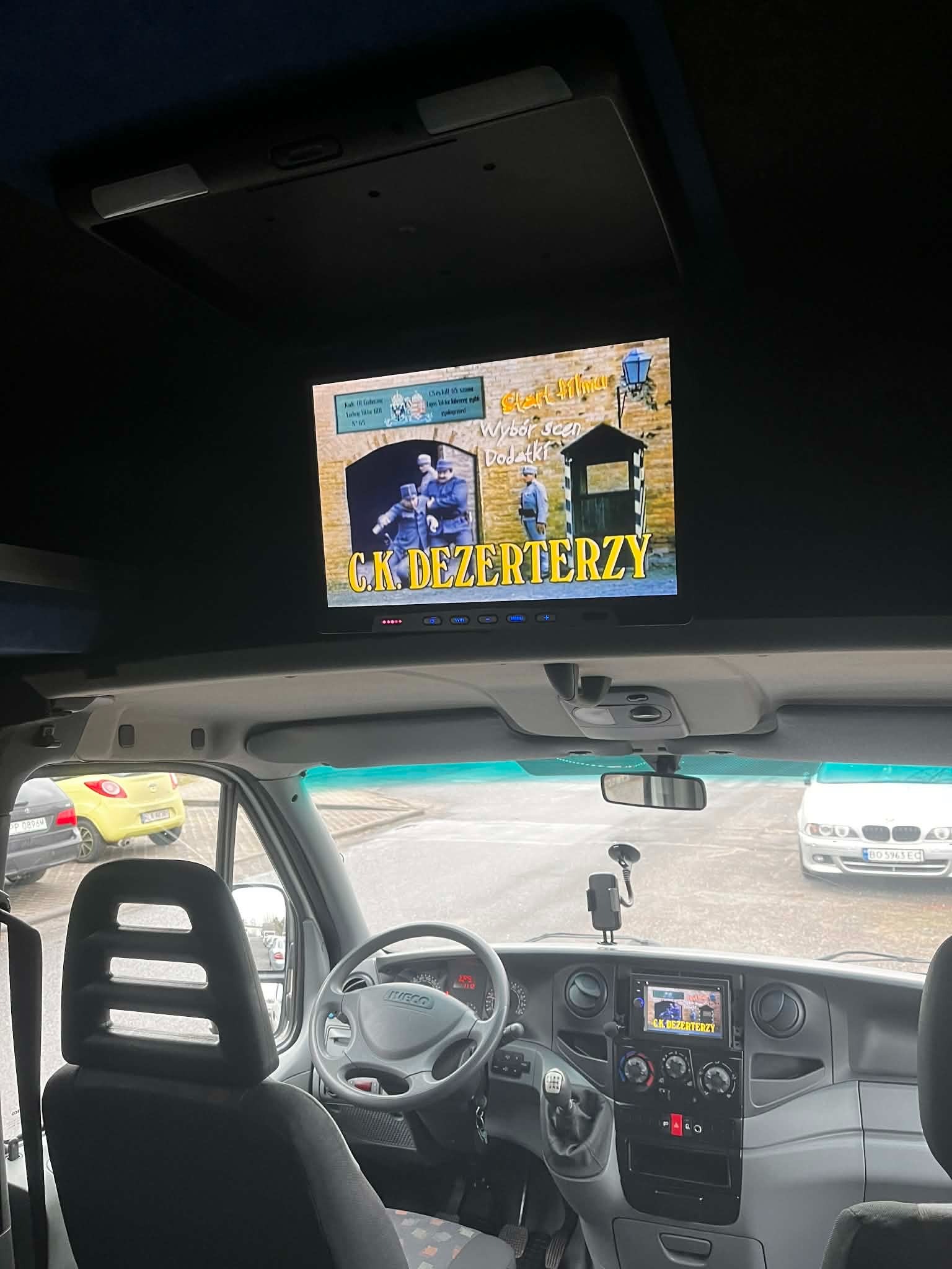 Wnętrze busa z ekranem sufitowym wyświetlającym film 'C.K. Dezerterzy', widok zza fotela kierowcy, deska rozdzielcza z radiem i widok na ulicę przez przednią szybę.