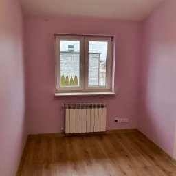 IM BAU sp. z o.o. - Pokój po malowaniu ścian na delikatny róż, z drewnianą podłogą i białym grzejnikiem pod oknem. Widok na zewnątrz przez okno.