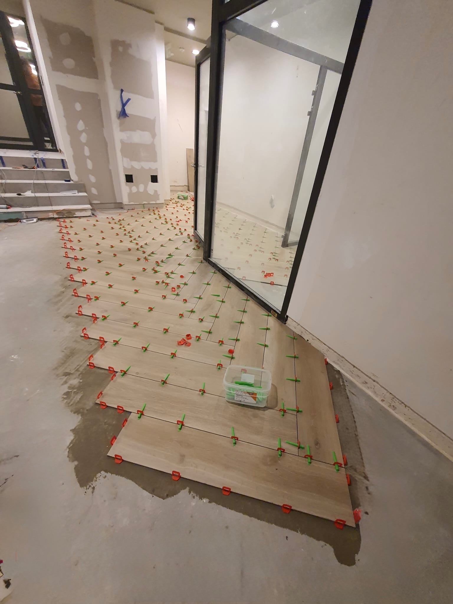 Układanie podłogi z płytek imitujących drewno, z użyciem klipsów poziomujących. Widoczne schody w tle i narzędzia glazurnicze. Praca w toku, jasne wnętrze.