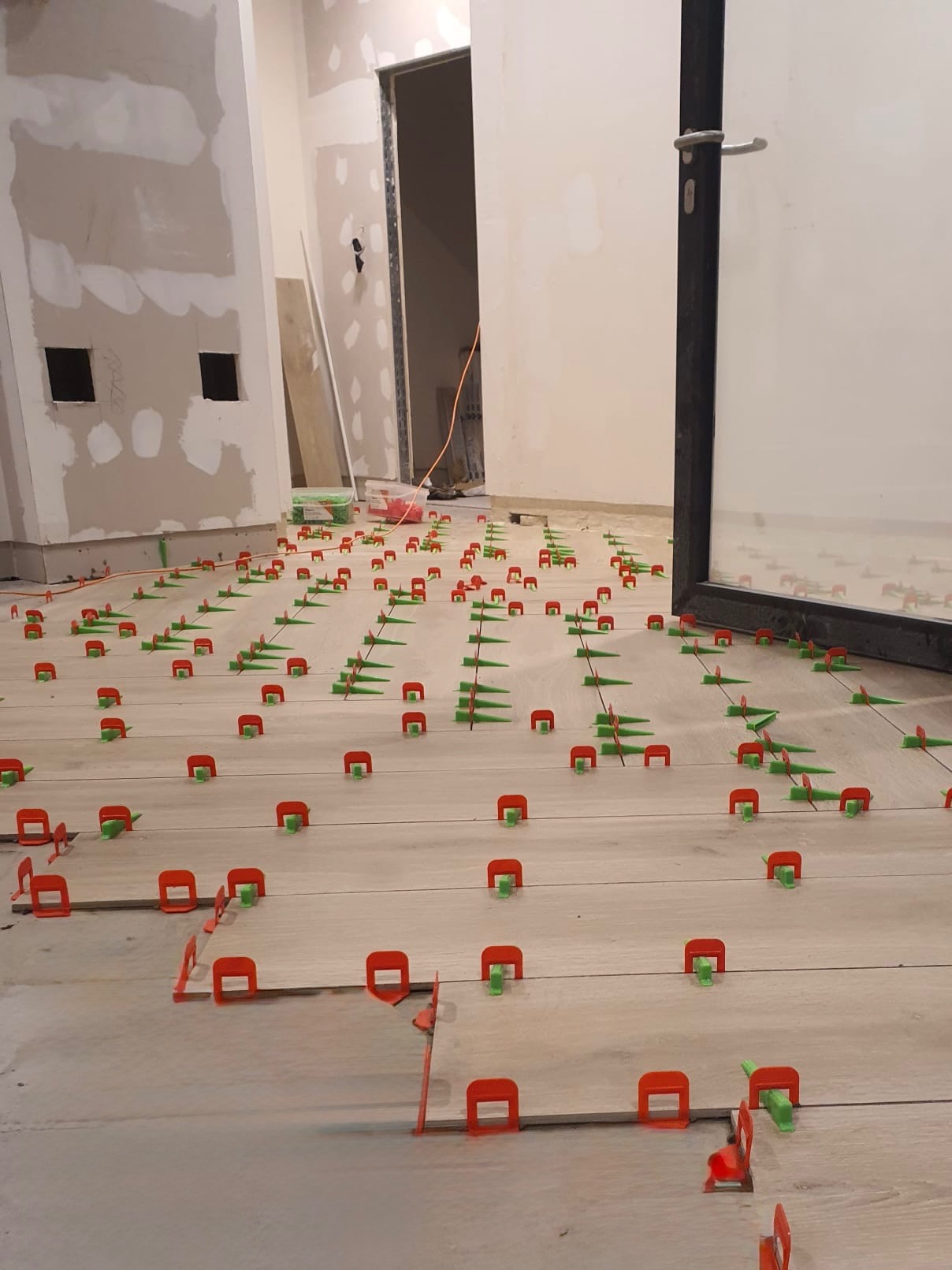 Układanie jasnych płytek podłogowych z użyciem czerwonych i zielonych klipsów poziomujących. W tle widoczne ściany przygotowane do szpachlowania i otwarte drzwi.