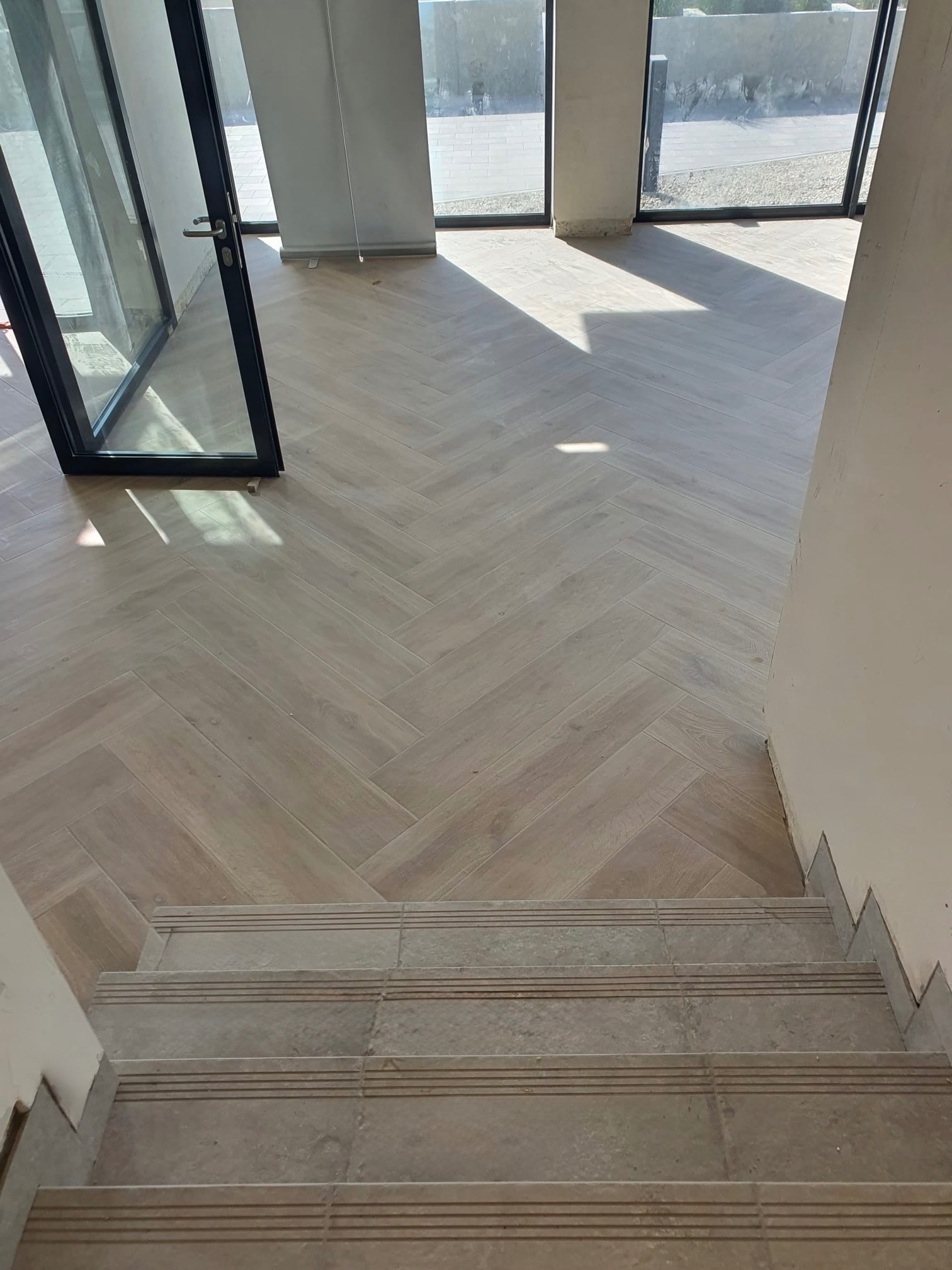 Ułożona jasna podłoga w jodełkę z płytek ceramicznych, widok z góry na schody z płytek, duże okna w tle w jasnym pomieszczeniu.