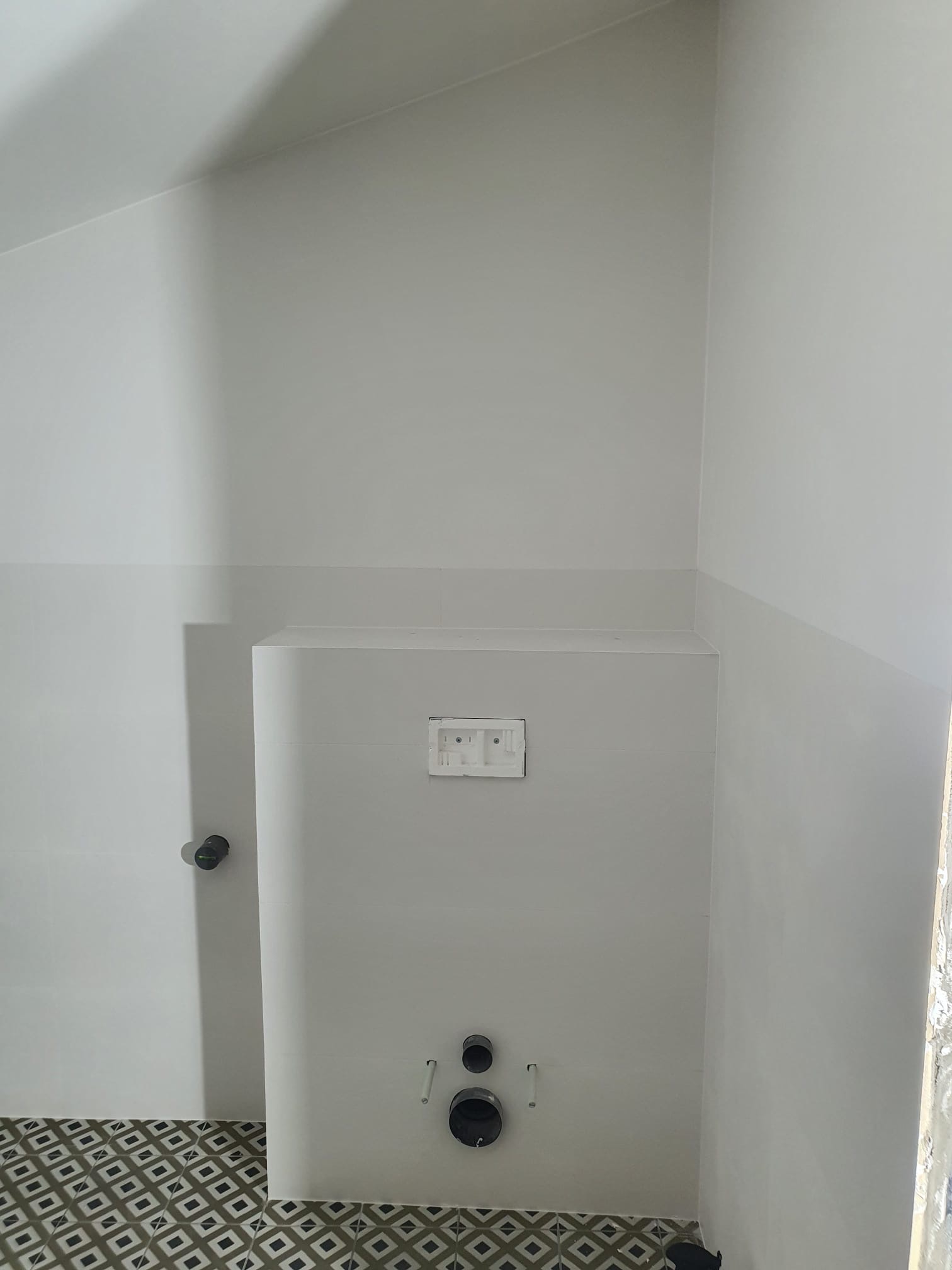 Wykończona łazienka z zabudowaną toaletą, białe ściany z szarym pasem płytek, wzorzysta podłoga. Widoczne przyłącza wodno-kanalizacyjne i przycisk spłuczki.