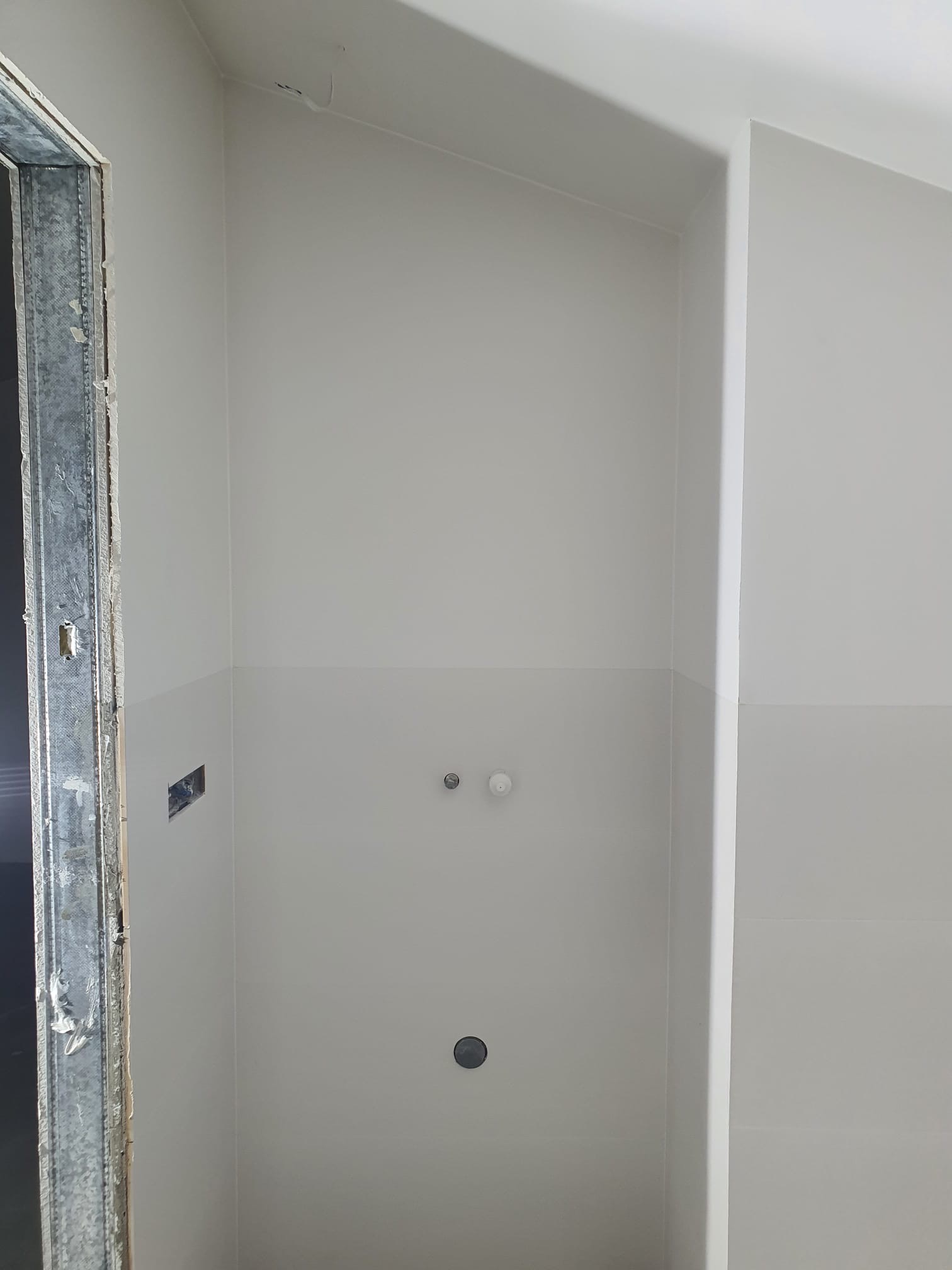 Wnętrze łazienki w trakcie prac wykończeniowych: ściany pomalowane na szaro, widoczne przyłącza wodne i metalowa ościeżnica drzwiowa przed montażem.