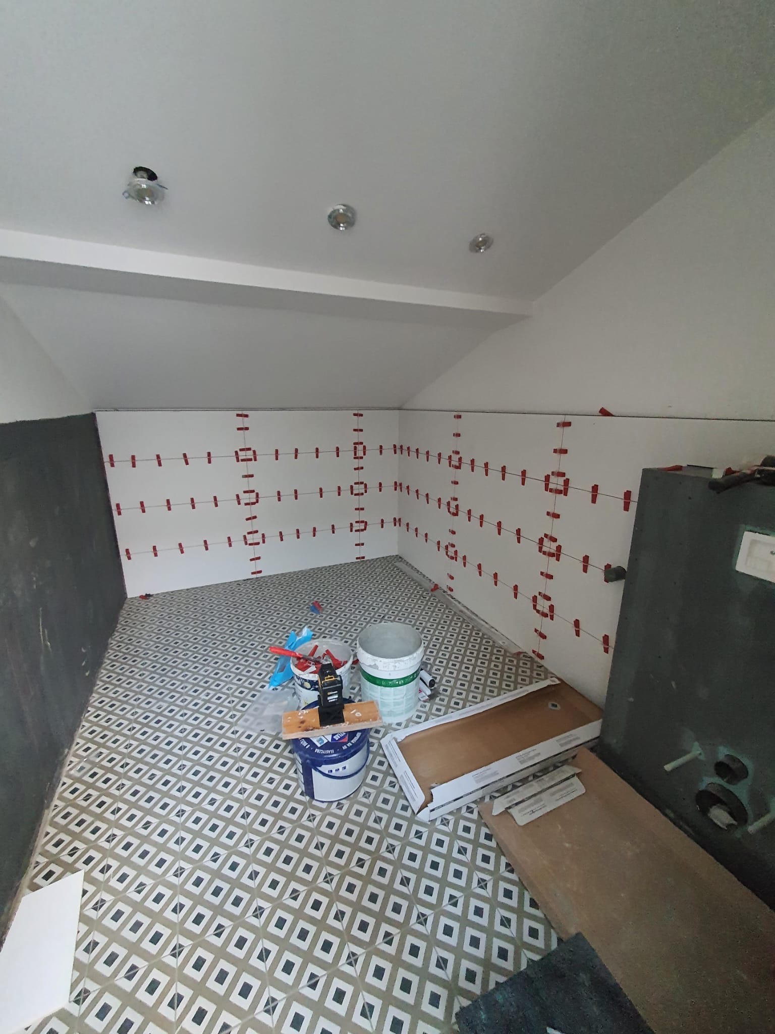 Układanie płytek w łazience na poddaszu. Widoczne poziomowanie płytek na ścianie za pomocą krzyżyków. Na podłodze wzorzyste płytki i narzędzia glazurnicze.