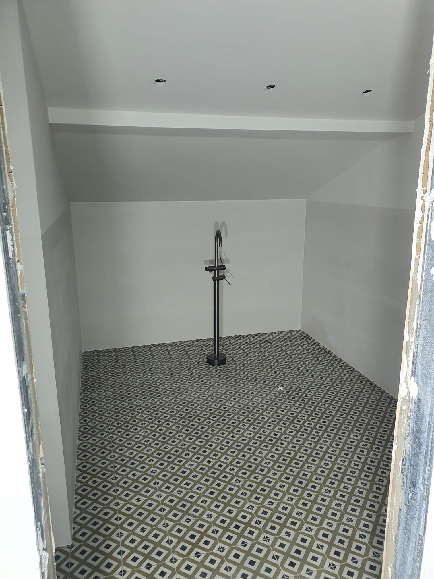 Wykończona łazienka z wzorzystą podłogą i czarną armaturą. Ściany pomalowane na biało, widoczne punkty świetlne w suficie. Ujęcie z progu, podkreślające przestrzeń.