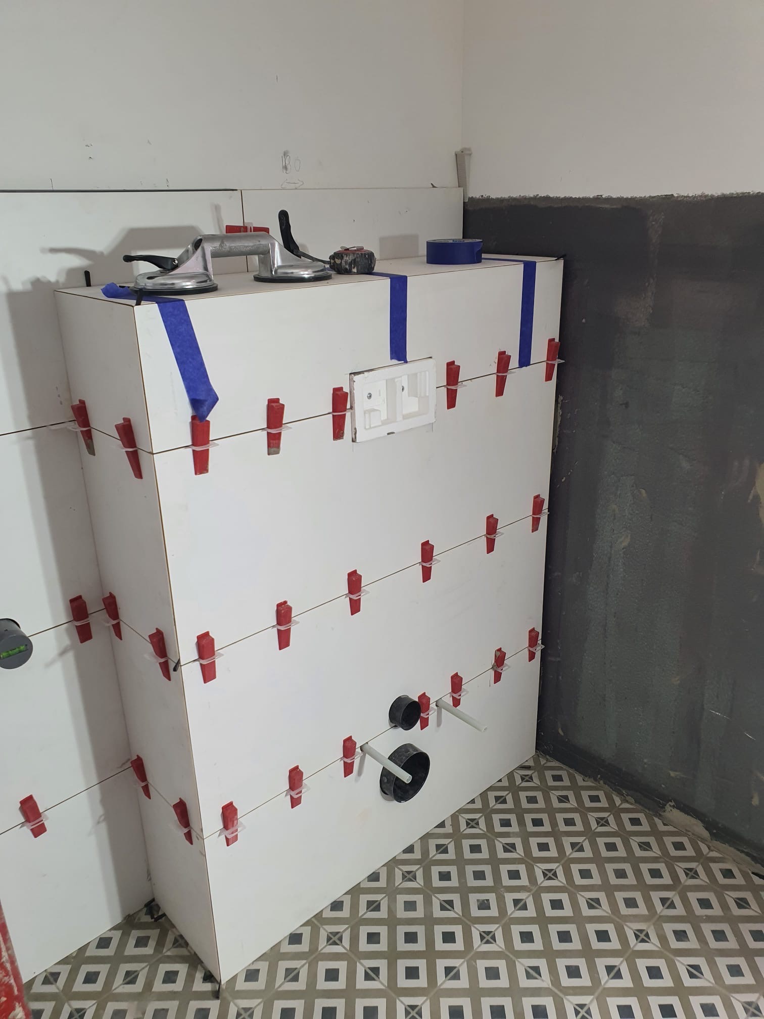 Biała obudowa stelaża WC w trakcie montażu płytek, z użyciem klipsów poziomujących i taśmy. Widoczne narzędzia glazurnicze i fragment wykończonej ściany. Wzorzysta podłoga.