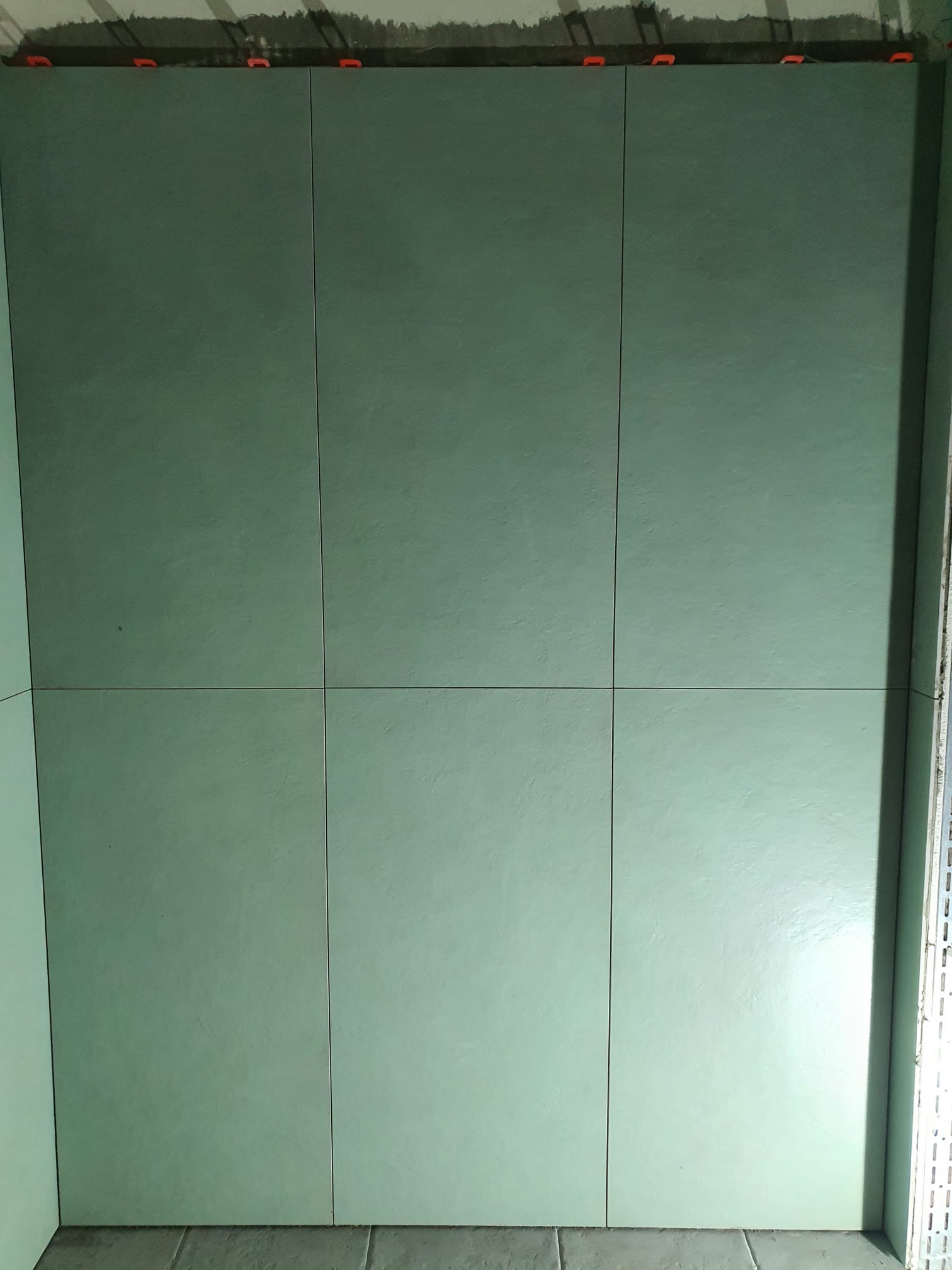 Ściana wyłożona dużymi, kwadratowymi, zielonymi płytkami. Widoczne pomarańczowe klipsy poziomujące na górnej krawędzi. Minimalistyczny design, świeżo położona glazura.