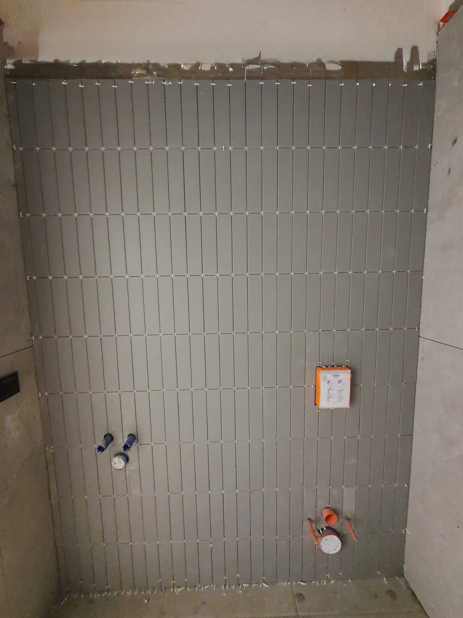 Ściana w trakcie układania szarych, podłużnych płytek w łazience. Widoczne krzyżyki dystansowe, instalacja wodna i fragment puszki elektrycznej.