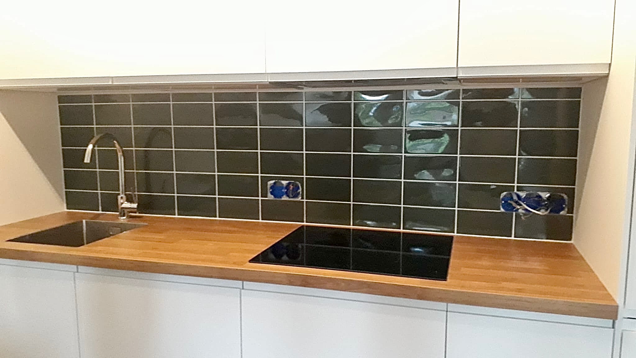 Nowoczesna kuchnia z ciemnozielonymi, błyszczącymi płytkami nad drewnianym blatem i białą zabudową. Widoczne gniazdka elektryczne przed montażem osprzętu.