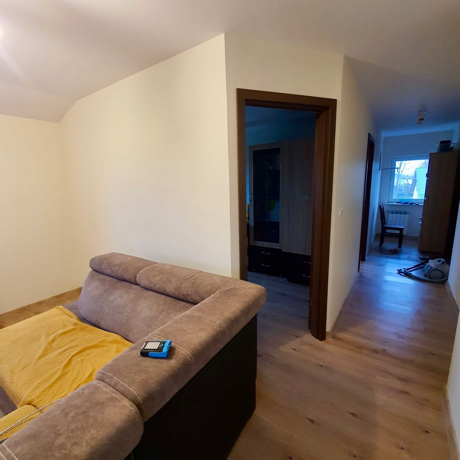 Jasne wnętrze po odświeżeniu: kremowe ściany, drewniana podłoga, widok na pokój z oknem. Sofa z żółtą narzutą w rogu kadru. Widoczny odkurzacz w tle.