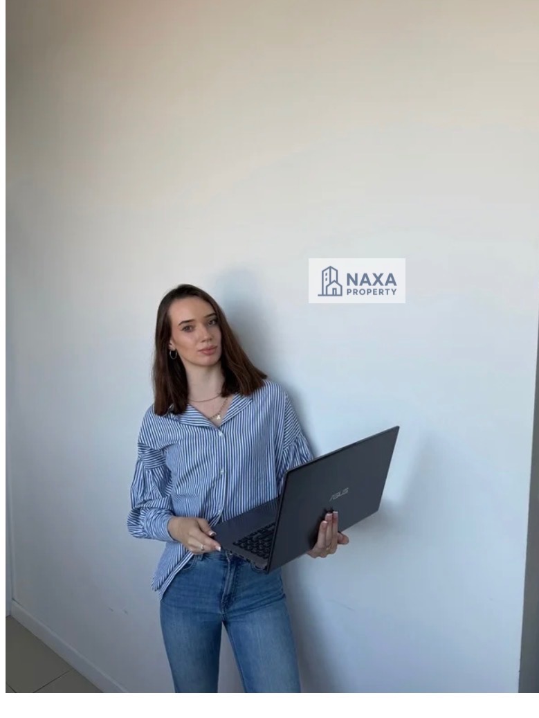 Kobieta w niebieskiej koszuli w paski trzyma laptopa na tle białej ściany. Widoczne logo firmy Naxa Property. Ujęcie od pasa w górę, naturalne światło.