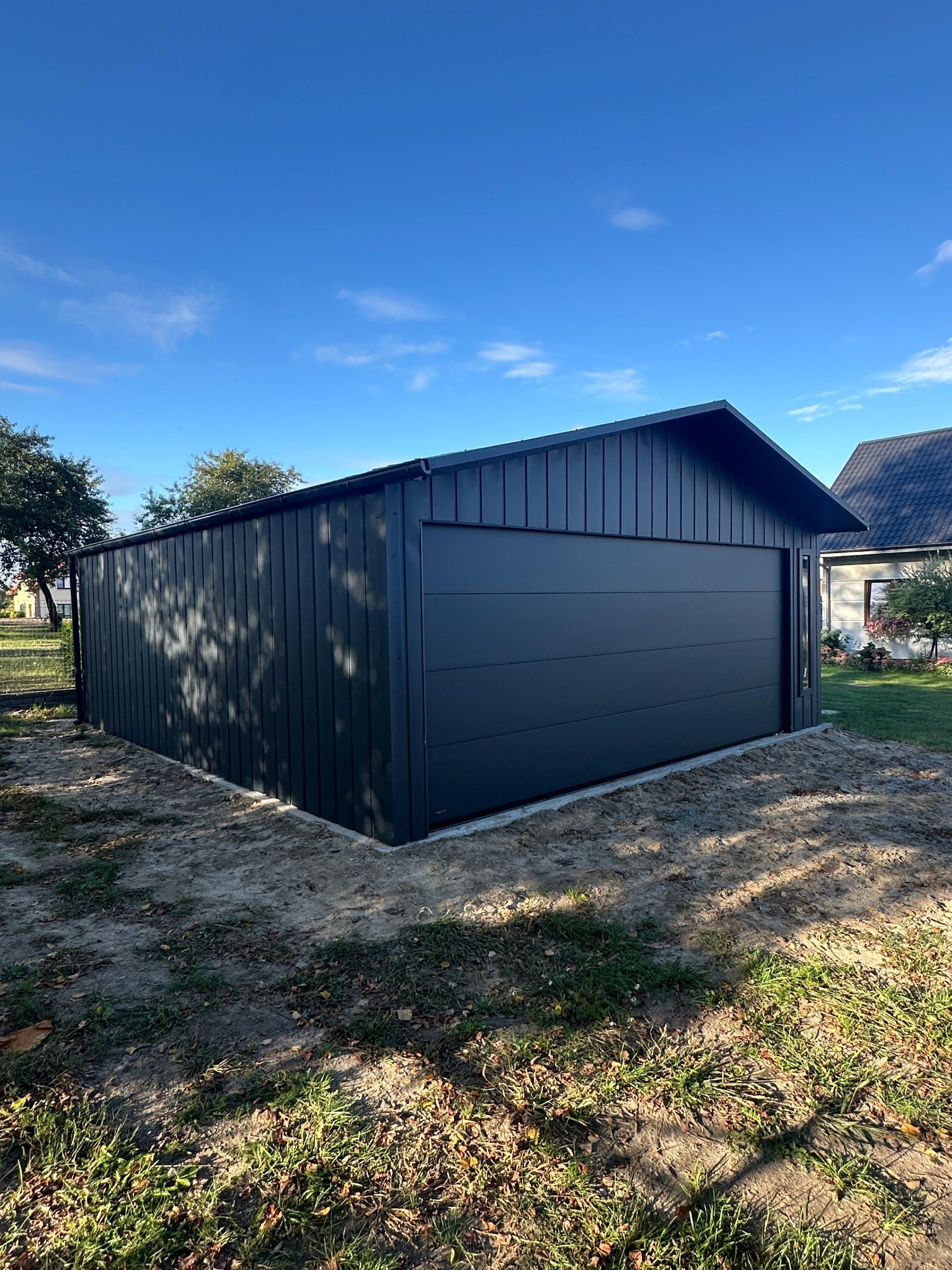 Czarny, matowy garaż blaszany z bramą segmentową, o wymiarach 6m x 5.80m, na tle błękitnego nieba i zieleni. Widok z zewnątrz, prezentujący gotowy produkt.