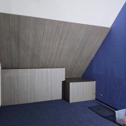 KSD Design - Sylwia Szczerkowska - Szafa wnękowa z szarego drewna pod skosem dachu, na niebieskiej wykładzinie. Widoczne elementy montażowe i narzędzia. Nietypowy układ szafy.
