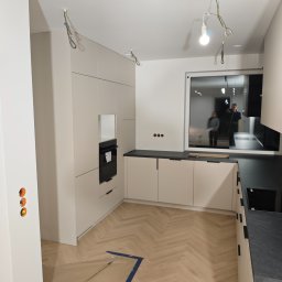 KSD Design - Sylwia Szczerkowska - Jasna kuchnia z czarnym blatem i sprzętem AGD w zabudowie. Na podłodze parkiet ułożony w jodełkę. Widoczne niedokończone prace elektryczne i zabezpieczenia podłogi.