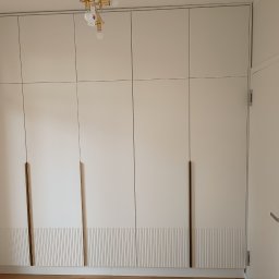 KSD Design - Sylwia Szczerkowska - Biała szafa wnękowa z frezowanymi frontami i długimi, smukłymi uchwytami w odcieniu złota, dopasowana do wysokości pomieszczenia, widok na fragment drzwi.